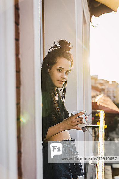 Portrait of beautiful young woman on balcony above the city at sunset with cup of tea