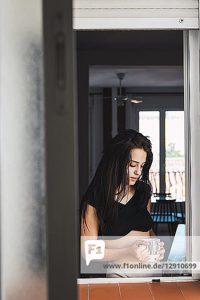 Serious beautiful young woman with cup of coffee at the window at home