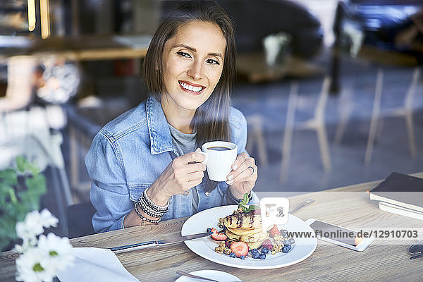 Portrait of smiling young woman enjoying pancakes and coffee in cafe