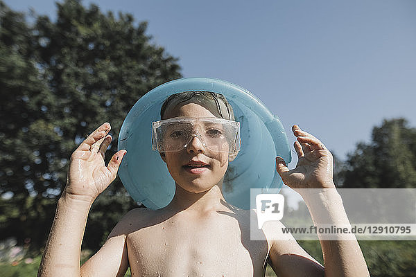 Wet boy wearing safety goggles holding bowl above his head in garden