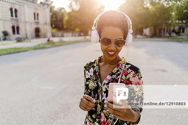 Happy fashionable young woman with headphones and smartphone outdoors at sunset