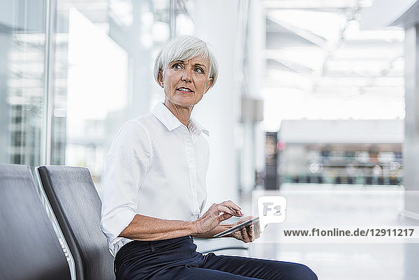 Senior businesswoman sitting in waiting area with tablet looking around