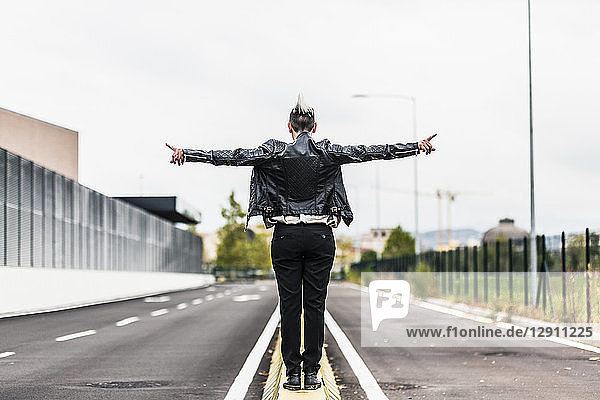 Rear view of punk woman standing on a barrier at the roadside with outstretched arms