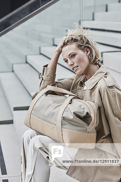 Portrait of blond businesswoman with travelling bag wearing beige trenchcoat sitting on stairs