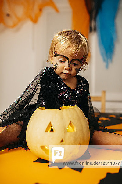 Portrait of little girl with painted face and fancy dress sitting on table playing with Jack O'Lantern