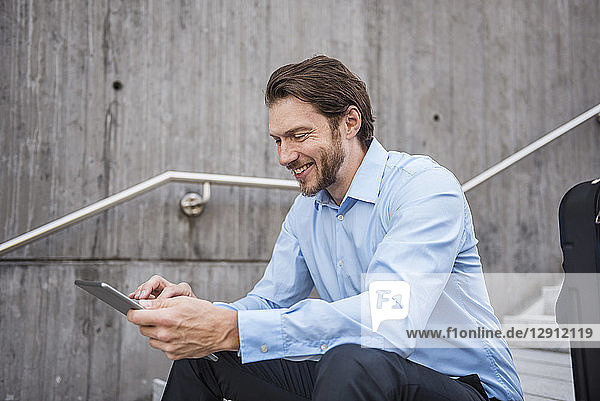 Smiling businessman with suitcase sitting on stairs using tablet