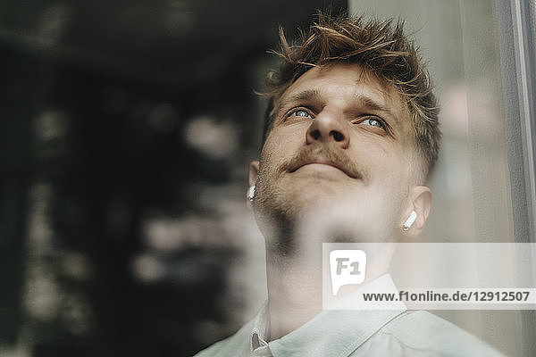 Young man standing at the window  using ear buds  daydreaming