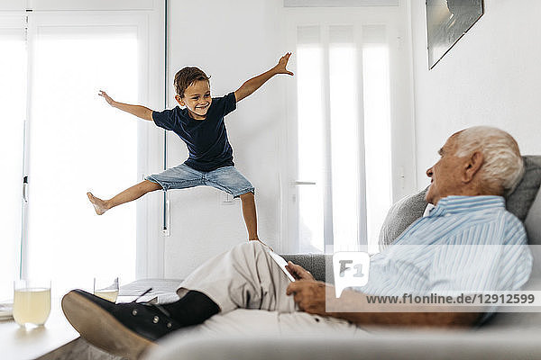 Grandfather sitting on the sofa with tablet watching his grandson jumping in the air