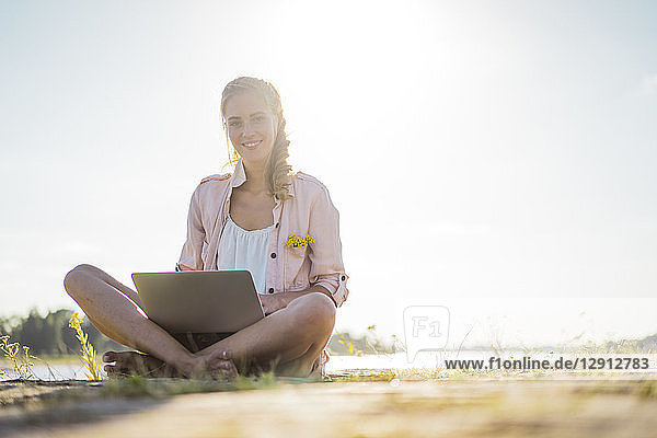 Portrait of smiling woman sitting at the riverside in summer using laptop