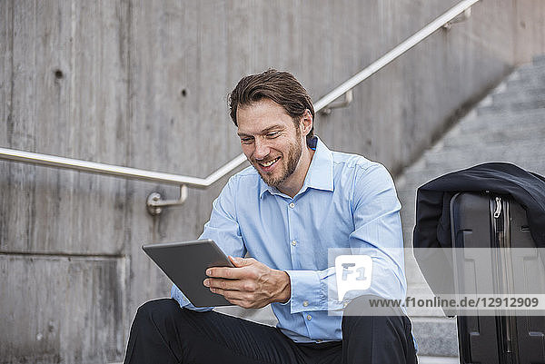 Smiling businessman with rolling suitcase sitting on stairs using tablet