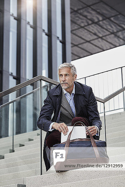 Portrait of mature businessman with traveeling bag and tablet crouching on stairs outdoors