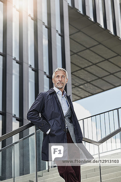 Portrait of mature businessman standing on stairs in front of modern office building looking at distance