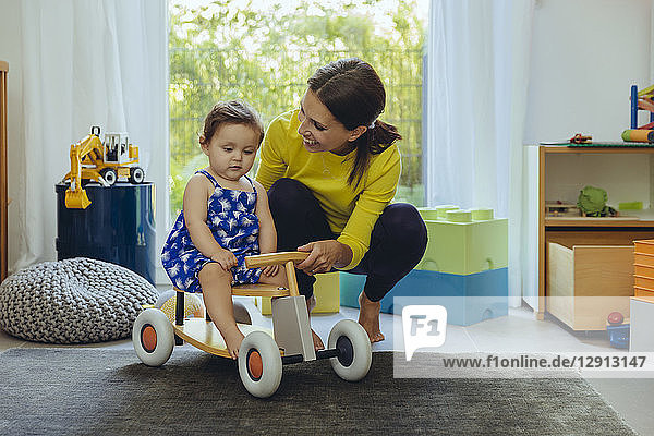 Happy mother with baby daughter on toy car in living room