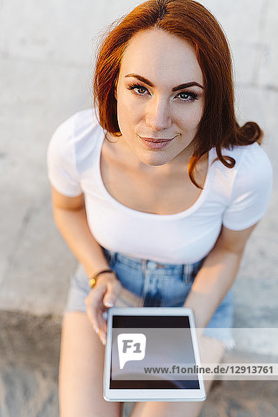 Portrait of smiling redheaded woman sitting on wall with digital tablet