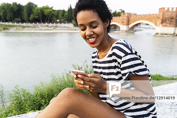 Smiling young woman sitting on wall at the riverside using cell phone