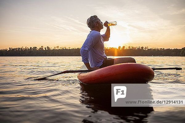Man sitting on paddleboard on a lake by sunset drinking beverage