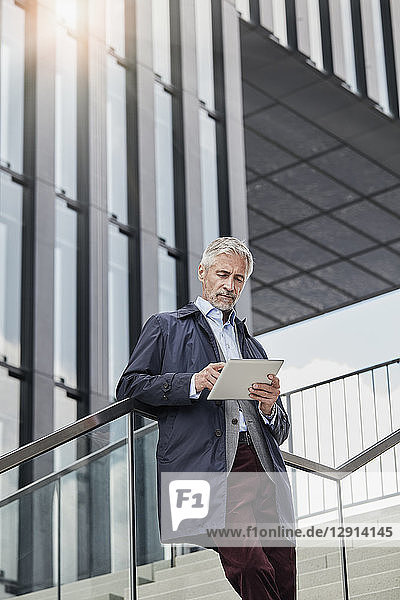 Portrait of mature businessman standing on stairs in front of modern office building using tablet