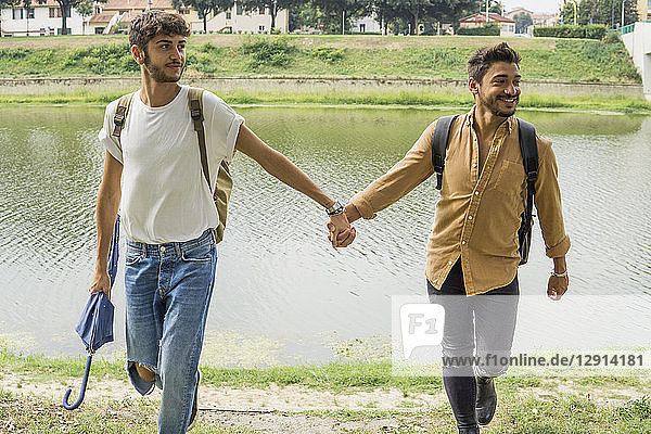 Portrait of young gay couple with backpacks walking hand in hand at riverside