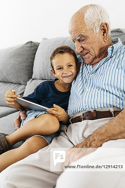Portrait of happy little boy with digital tablet sitting besides his grandfather on the couch at home