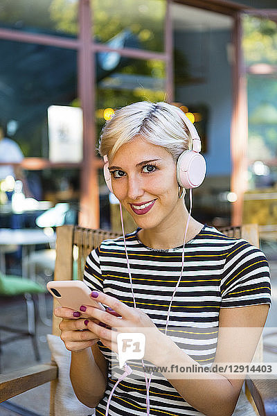 Portrait of smiling young woman with smartphone listening music with headphones at pavement cafe