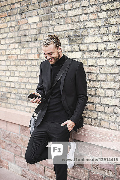 Smiling fashionable young man leaning against brick wall using cell phone