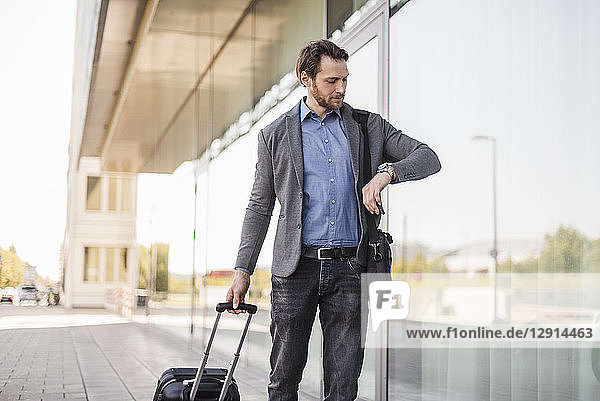 Businessman with rolling suitcase and laptop bag checking the time