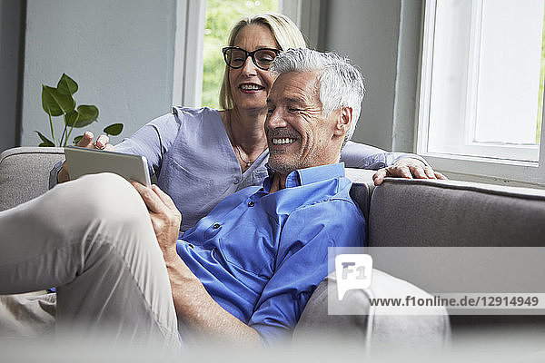 Happy mature couple sitting on couch at home sharing tablet