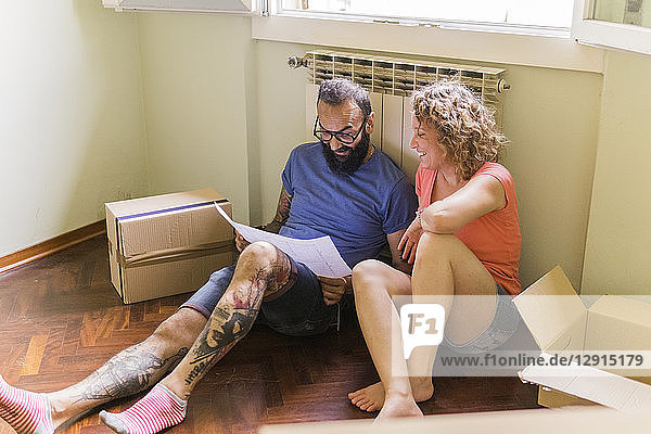 Happy couple sitting side by side on the floor of new home looking at plan