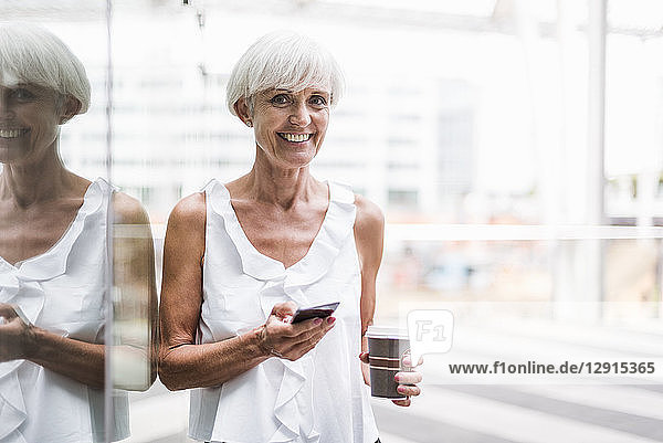 Portrait of smiling senior woman with cell phone and takeway coffee outdoors