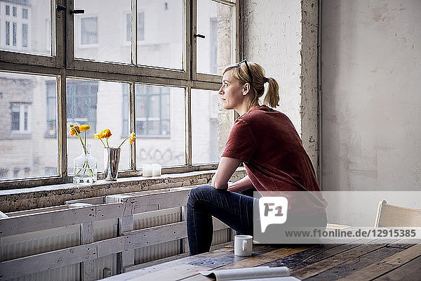 Woman sitting on desk in loft looking through window