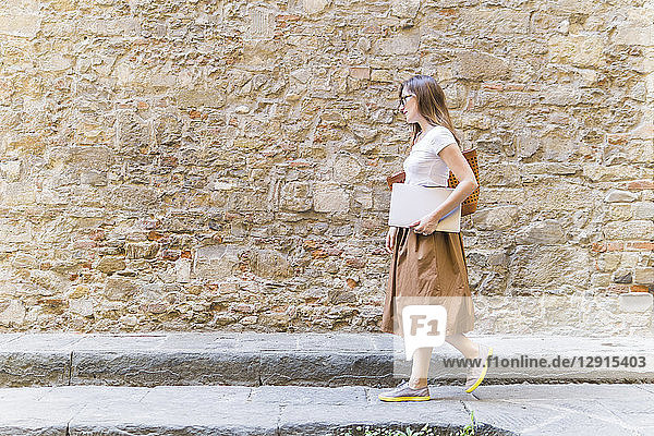 Woman walking along a stone wall carrying laptop