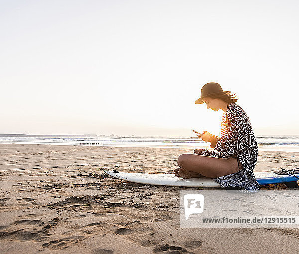 Young woman sitting on surfboard at the beach  using smartphone