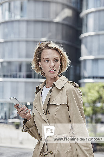 Germany  Duesseldorf  portrait of blond young businesswoman with smartphone wearing beige trenchcoat