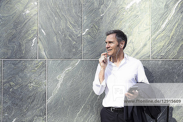 Mature businessman with earphones and smartphone at a wall
