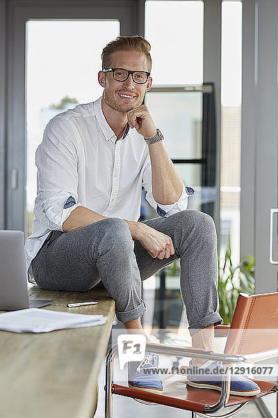 Portrait of smiling businessman with laptop sitting on desk in office