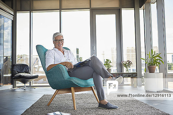 Portrait of smiling mature man with a brochure relaxing in armchair at the window at home
