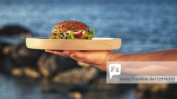 Woman's hand holding wooden plate with fishburger in front of the sea