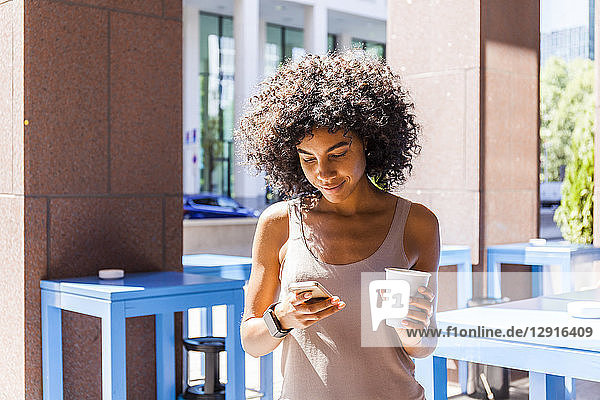 Smiling young woman with coffee to go looking at cell phone