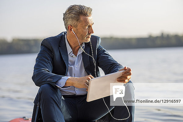 Mature businessman with earphones and tablet relaxing at lake