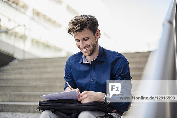 Smiling businessman sitting on stairs in the city taking notes