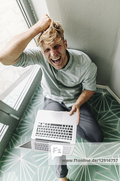 Young man sitting on ground  holding laptop  screaming in despair