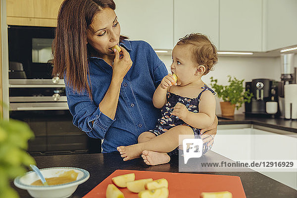 Mother and baby daughter eating apple chunks in kitchen together