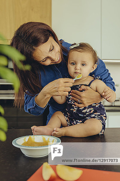 Mother helping baby daughter eating fruit pulp in kitchen