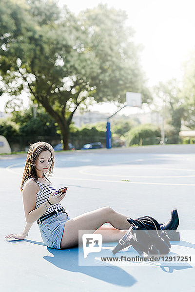 Young woman with backpack sitting on sports ground using cell phone