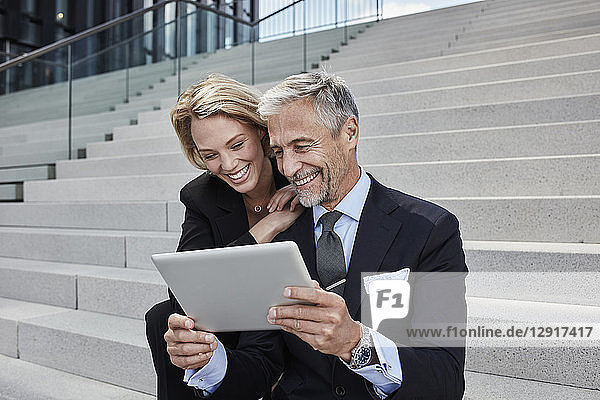 Portrait of two laughing businesspeople sitting together on stairs looking at tablet
