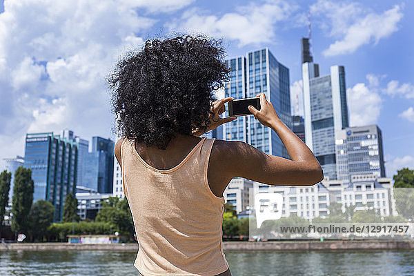 Germany  Frankfurt  back view of young woman with curly hair taking photo with smartphone