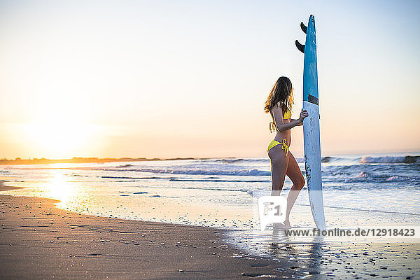Side view and full length shot of woman in yellow bikini with surfboard on beach