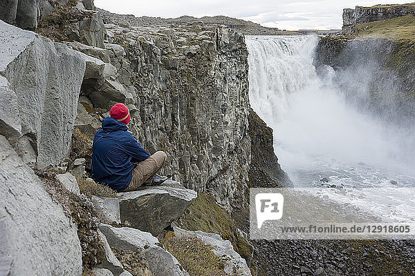 Ein junger Mann mit blauer Jacke und roter Mütze setzt sich auf einen Felsen an der Steilwand und bewundert den Dettifoss-Wasserfall in Nordisland.