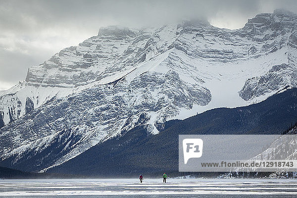 Fernblick auf zwei Personen beim Schlittschuhlaufen auf dem zugefrorenen Lake Minnewanka im Winter  Banff National Park  Alberta  Kanada