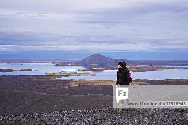 A young woman dressed in black and wearing boots walks along the ridge of the Hverfjall crater with Lake Myvatn in the background.